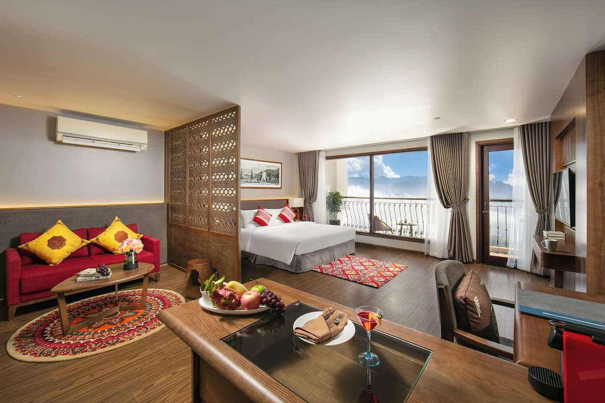 Habitación de hotel con cama, zona de estar con sofá rojo, mesa de comedor con fruta y una bebida, y balcón con vistas.