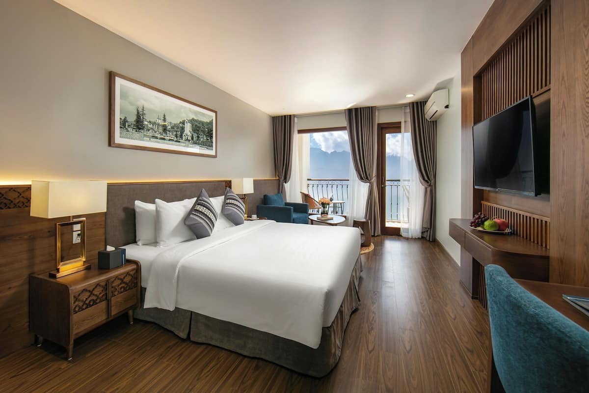 Habitación de hotel con una cama grande, televisión, escritorio con silla, balcón pequeño y un cuadro en la pared.