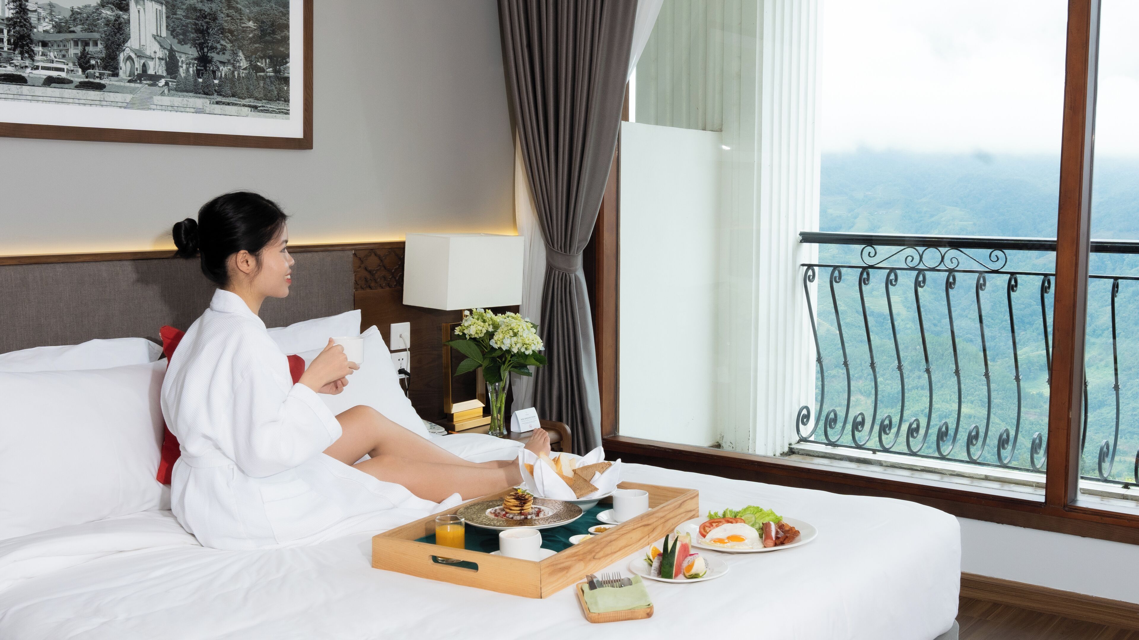 Een hotelkamer met een bed, een ontbijtplateau, een vaas met bloemen en uitzicht op zee vanaf een balkon.