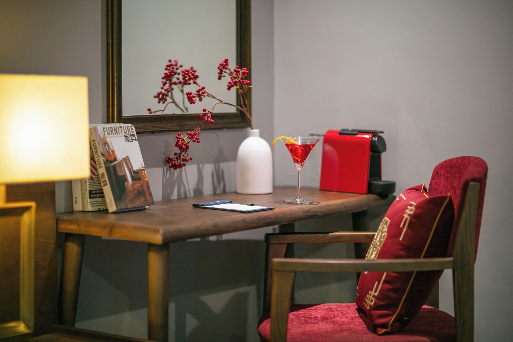 A wooden table with a magazine, a martini glass with a garnish, a white bottle, and a red suitcase.