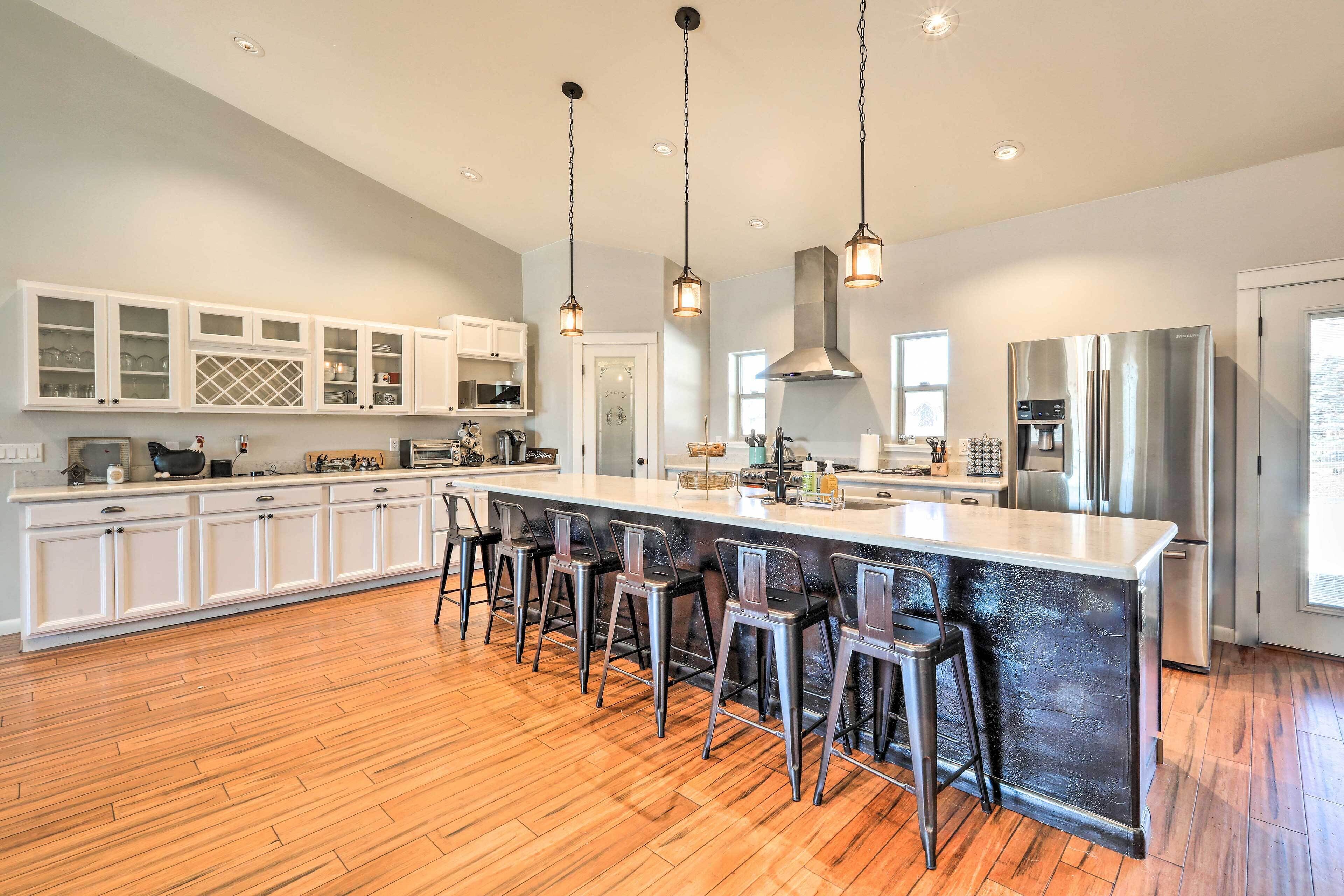 Una cocina moderna con una isla grande, taburetes de bar, muebles blancos, electrodomésticos de acero inoxidable e iluminación colgante.