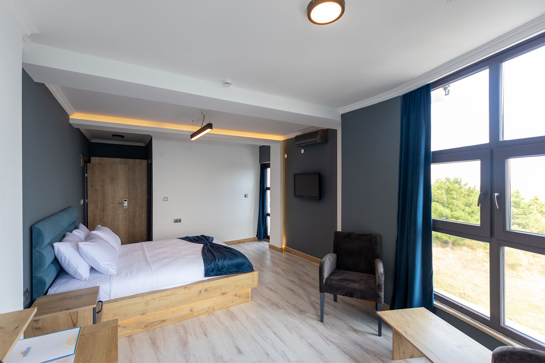 A modern hotel room with a bed, a desk, a chair, and a large window offering a view of the outdoors.