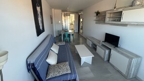 Uma sala de estar com sofá, televisão, mesa de jantar com cadeiras e área de cozinha ao fundo.