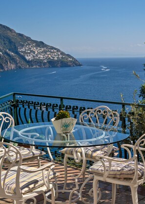 A glass table with chairs and a potted plant on a balcony overlooking the sea.