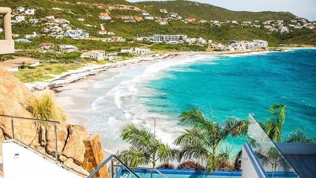 A coastal hotel with a pool overlooking turquoise waters, a staircase leading to the pool, and a view of the beach and hillside town. - Villa Amalia – 6-Bedroom Luxury Oceanview Villa with Pool, Gym & Full Staff in St. Maarten (Philipsburg)