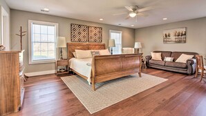 A bedroom with a wooden bed, two bedside tables with lamps, a brown leather sofa, and a large window with blinds. - Morrisonville Manor w/ Fire Pit & Hot Tub! (Lovettsville)