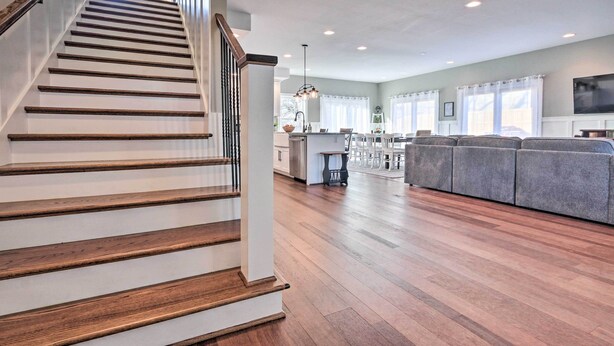 A spacious living area with a wooden staircase, a gray sofa, and a kitchen in the background. - Morrisonville Manor w/ Fire Pit & Hot Tub! (Lovettsville)