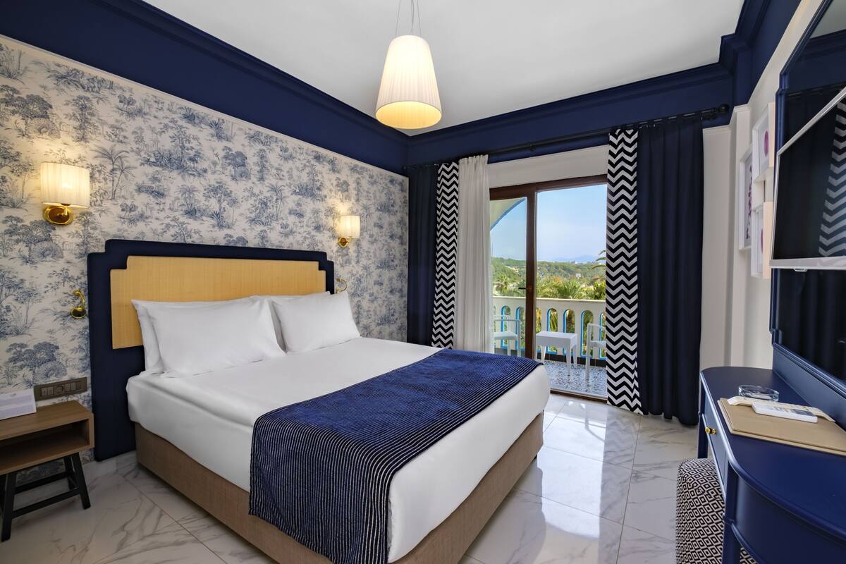 A hotel room with a large bed, a nightstand, a desk, a TV, and a balcony with a view.