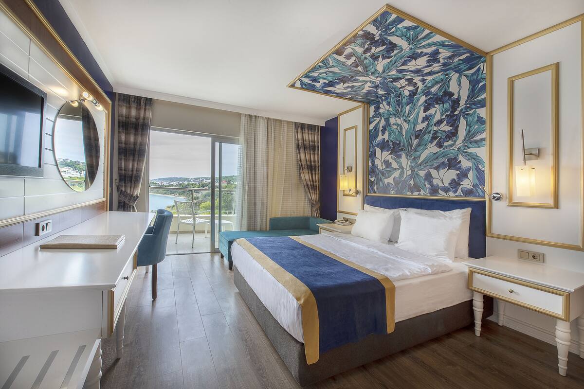 A hotel room with a large bed, a desk, a chair, a large window with a view, and a decorative wall.