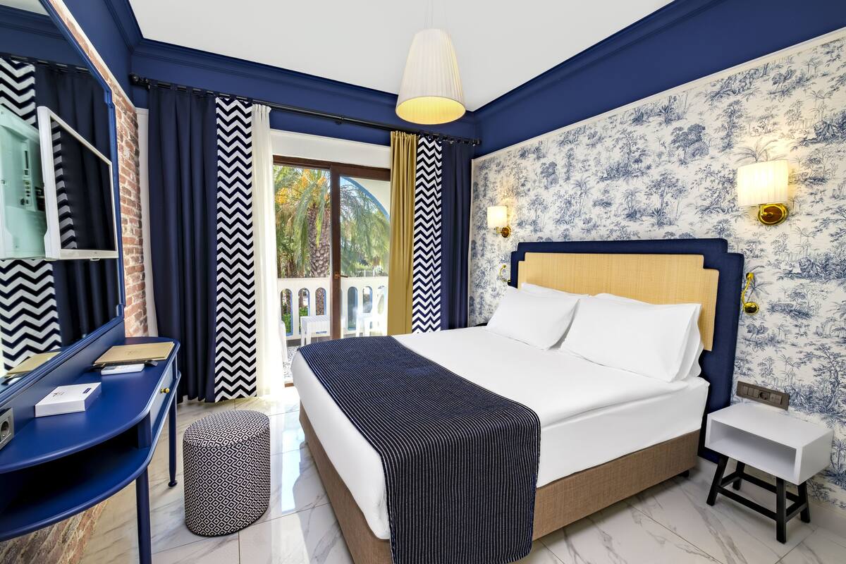 A hotel room with a bed, bedside table, lamp, and a view of the outdoors.