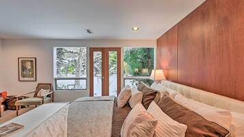 A modern bedroom with a large bed, a desk, and a chair. There are large windows with a view of greenery outside.