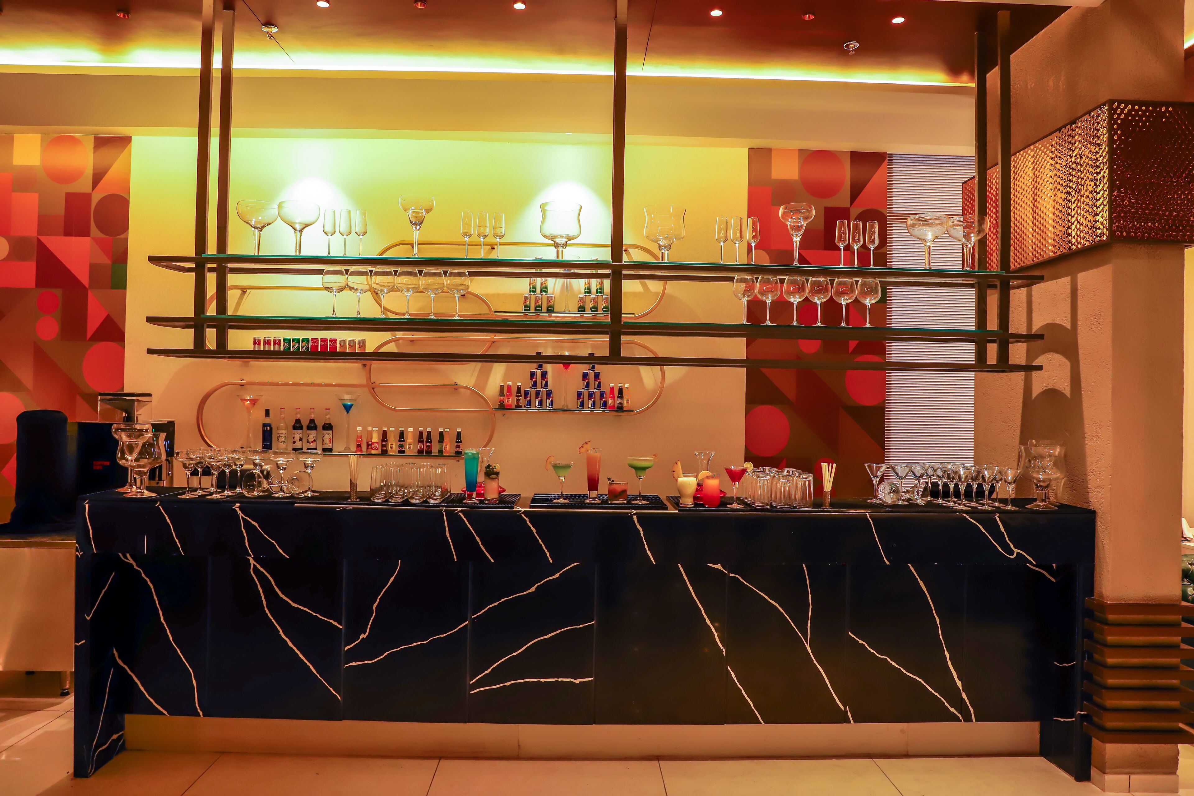 Eine Bar mit einer schwarzen Marmorplatte, Regalen voller Gläser und Flaschen sowie einer Wand mit geometrischem Muster.