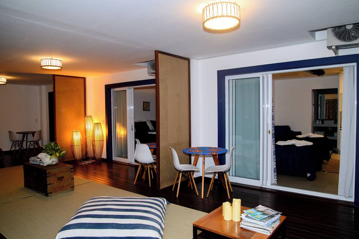 Habitación de hotel con cama, una mesita con revistas, una silla, una lámpara de mesa y una puerta corrediza de vidrio que da a un balcón.
