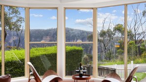 A room with large windows offering a view of a hilly landscape. There are two chairs and a small table with a bottle and glasses. - outstanding escarpment and valley views, close to lookouts and bushwalks (Katoomba)