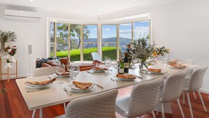 A dining area with a large table set for six, white chairs, and a view of greenery and hills through large windows. - outstanding escarpment and valley views, close to lookouts and bushwalks (Katoomba)