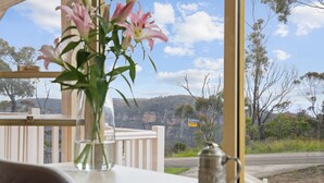 A table with a vase of flowers, a glass of water, and a metal container. A white railing and a road with a sign are visible through the window. - outstanding escarpment and valley views, close to lookouts and bushwalks (Katoomba)