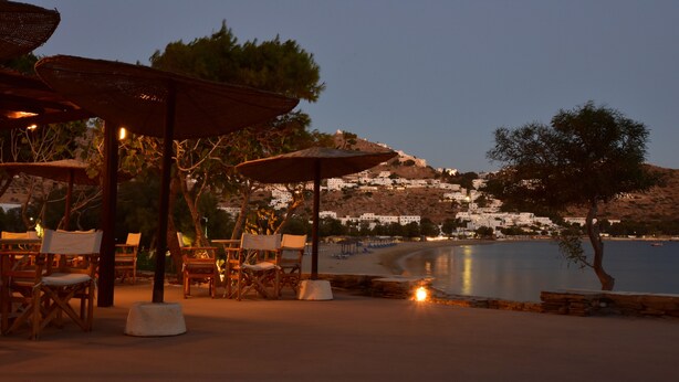 An outdoor seating area with wooden chairs and umbrellas, overlooking a beach with white buildings on a hillside. - Petra Holiday Village (Ios)