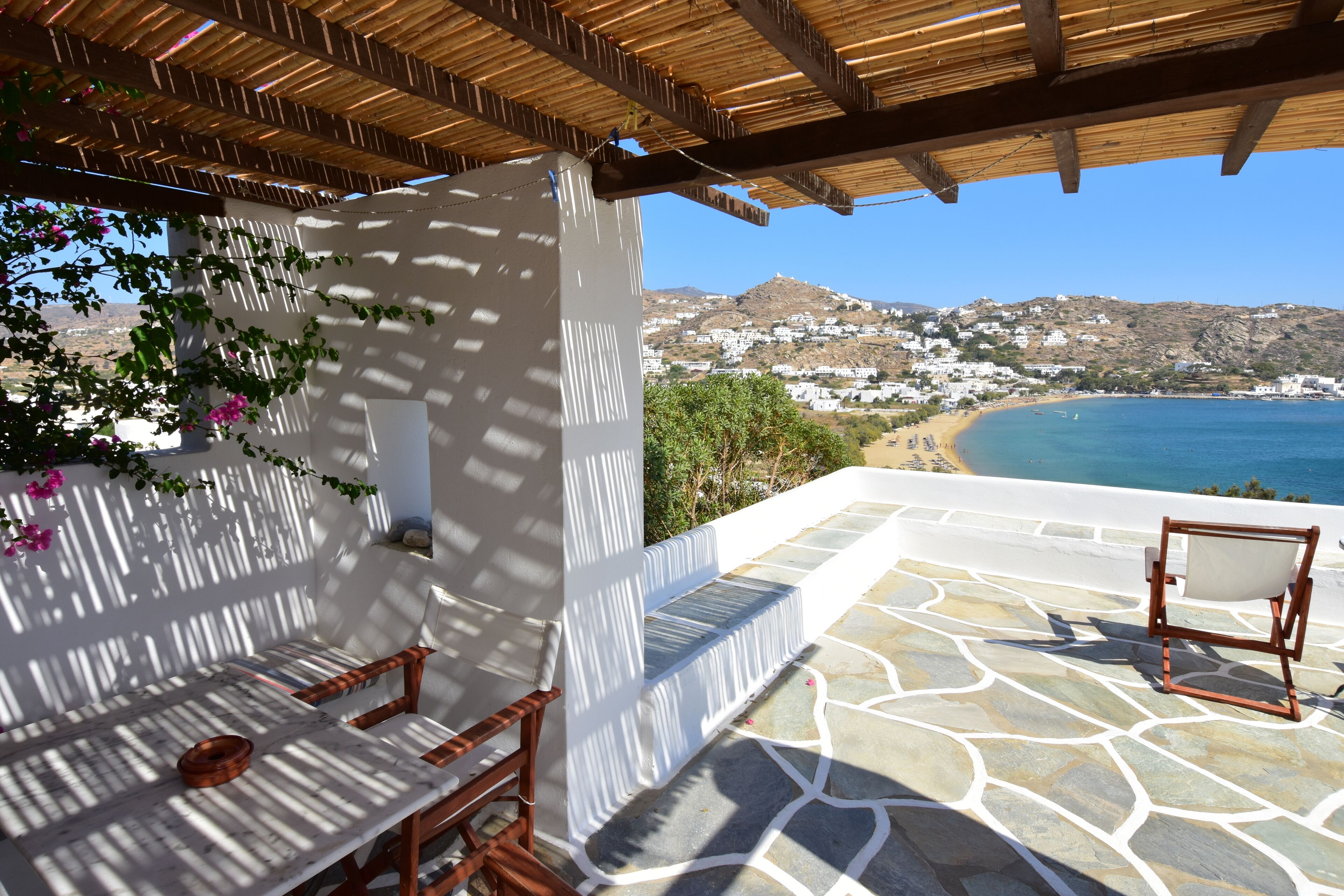 A terrace with stone flooring, white walls, and wooden furniture. A view of a beach and a hilly landscape is visible in the background.