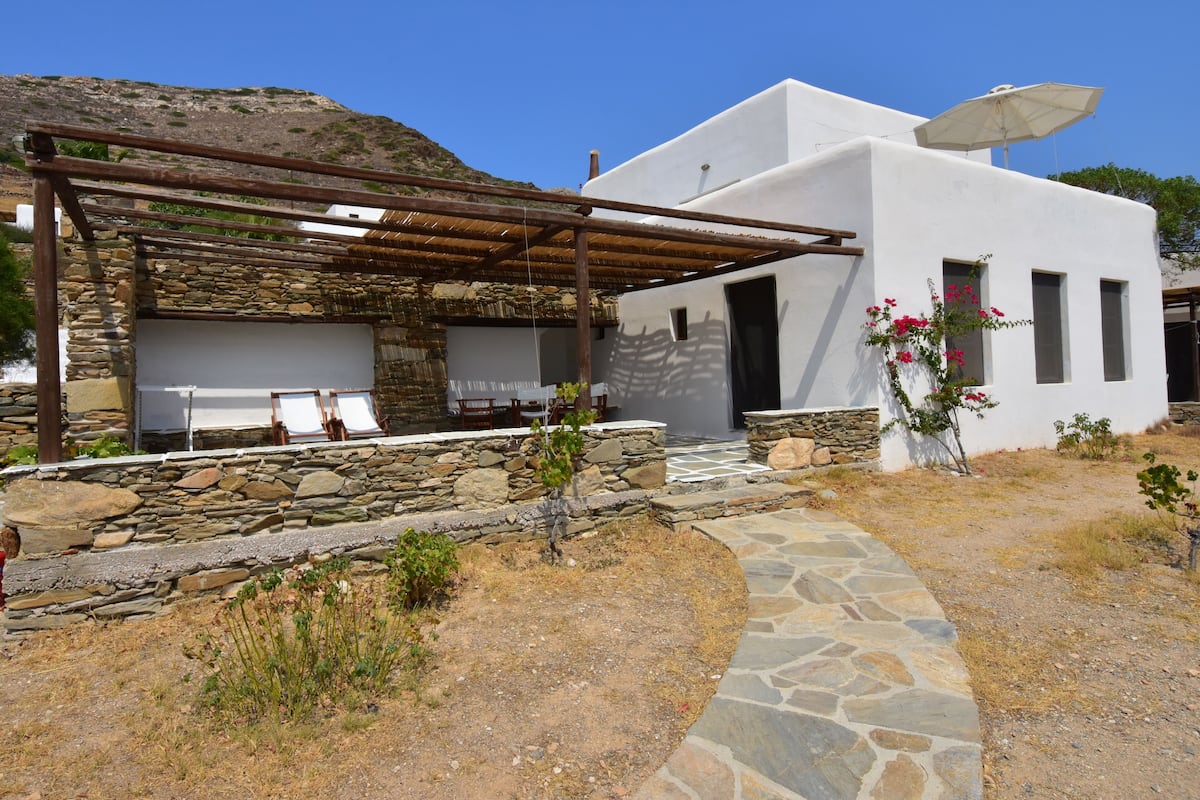 A white building with a stone exterior, a pergola with wooden beams, and a stone pathway leading to the entrance.