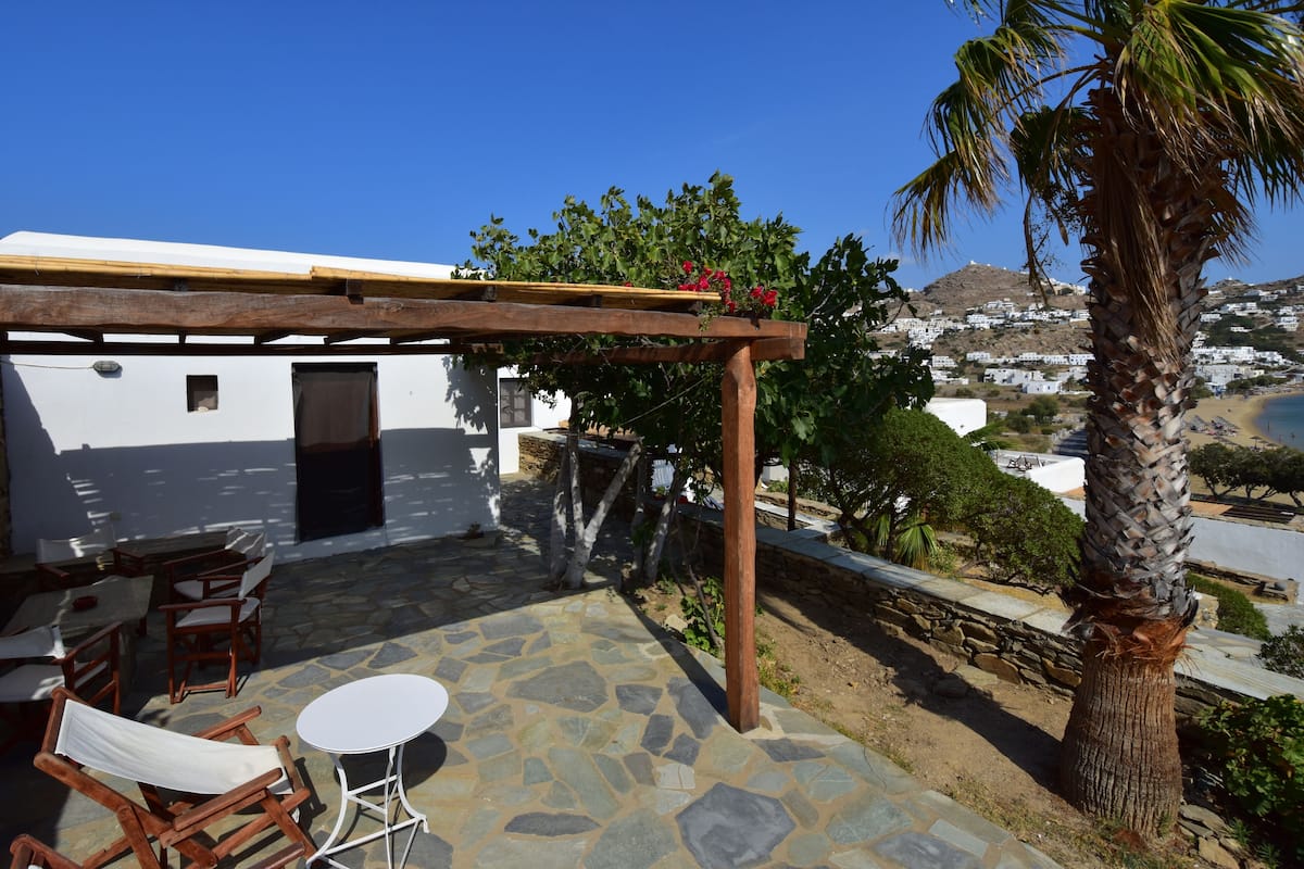 A covered outdoor seating area with stone flooring, wooden furniture, and a view of a coastal town.