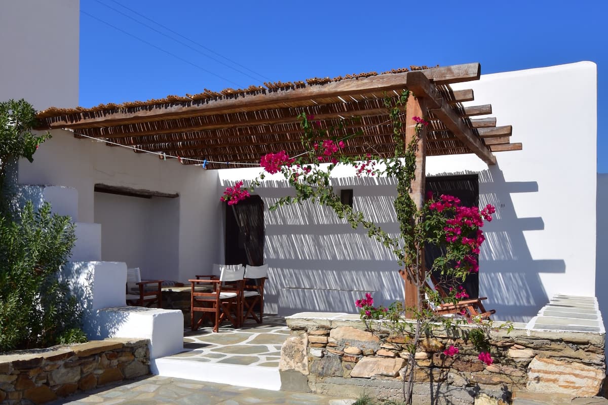 A stone-paved courtyard with a wooden pergola, a white wall, and a small table with chairs.