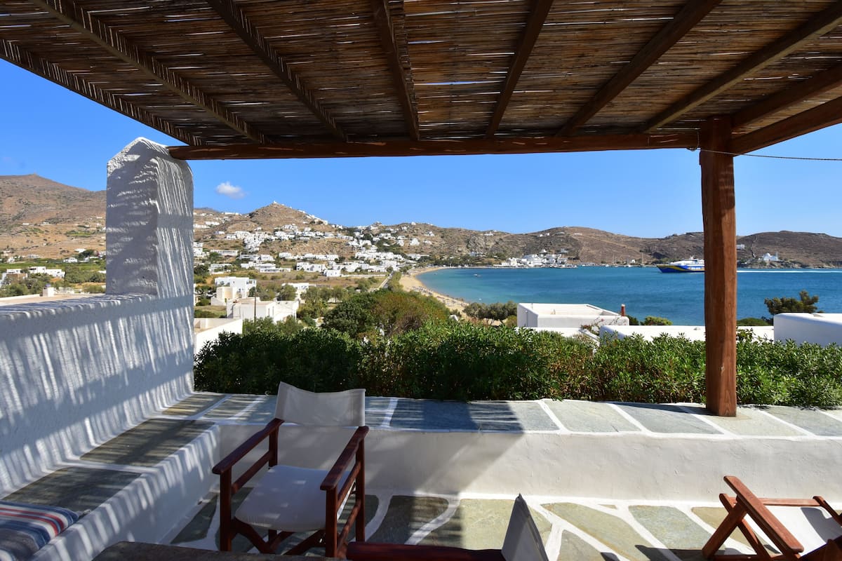 A covered outdoor seating area with stone flooring and wooden chairs, overlooking a coastal town with white buildings and a clear blue sea.