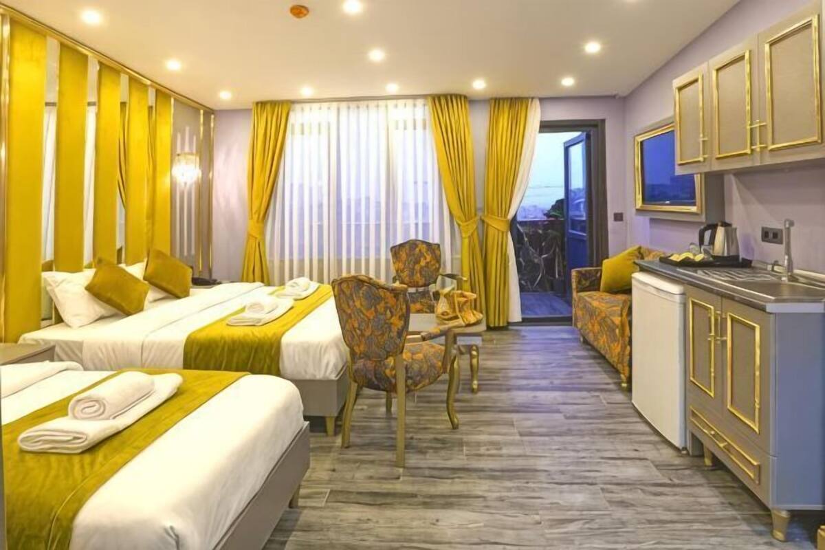 A hotel room with two beds, a dining table, a chair, a kitchen area, and a view through a door.