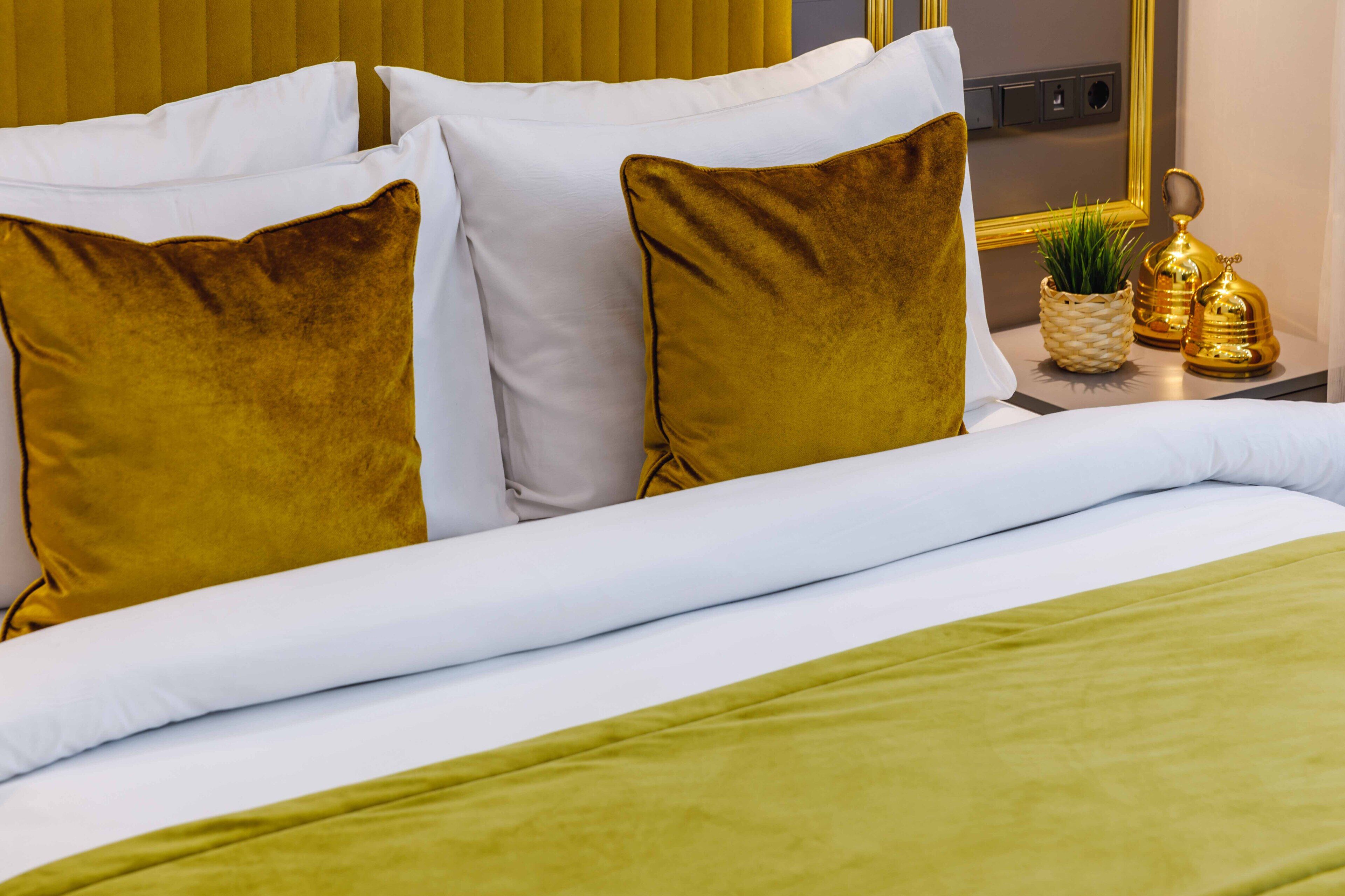 Una cama bien hecha con almohadillas blancas y doradas, una manta verde y una mesita con una planta y una campana dorada.