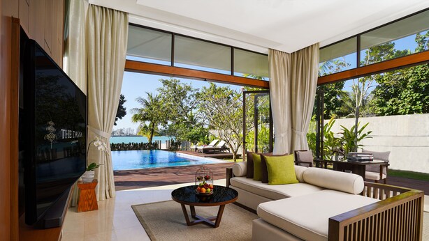 A modern living room with a large TV, a sofa, a coffee table with fruit, and a view of a swimming pool and greenery. - The Danna Beach Villas - A Member of Small Luxury Hotels of the World (Langkawi)