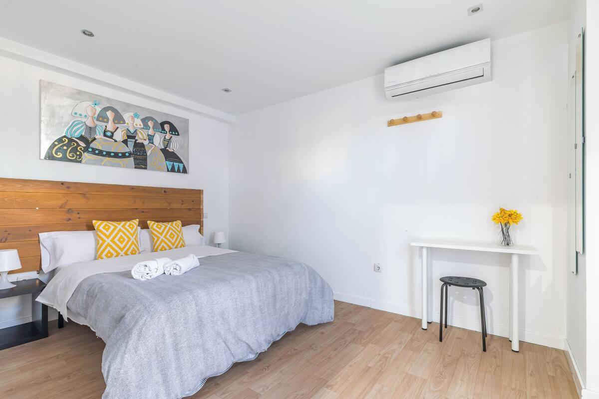 Una habitación de hotel moderna con cabecera de madera, una cama grande con edredón gris, una mesita blanca pequeña con un jarrón de flores amarillas y un aire acondicionado instalado en la pared.