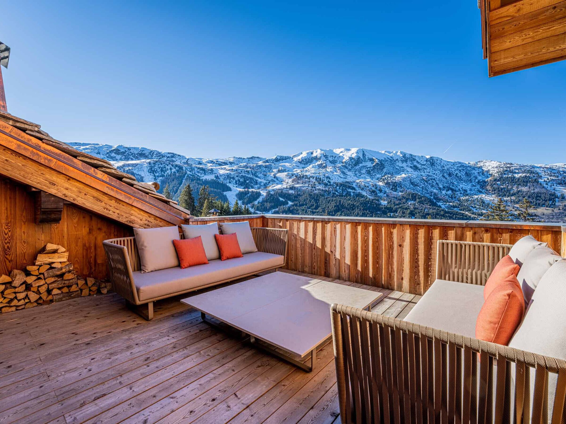 Une terrasse en bois avec un canapé et des fauteuils, une table et une pile de bois de chauffage.