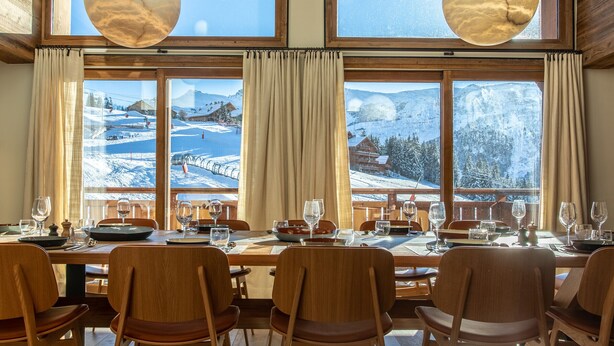 Zona pranzo con un grande tavolo apparecchiato per sei persone, ampie finestre con vista su una montagna innevata e travi in legno al soffitto.