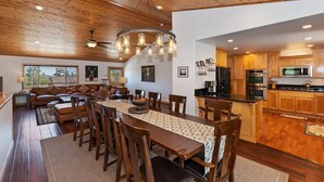 A spacious living area with a wooden ceiling, a dining table with chairs, a kitchen with wooden cabinets, and a large window offering a view of trees.