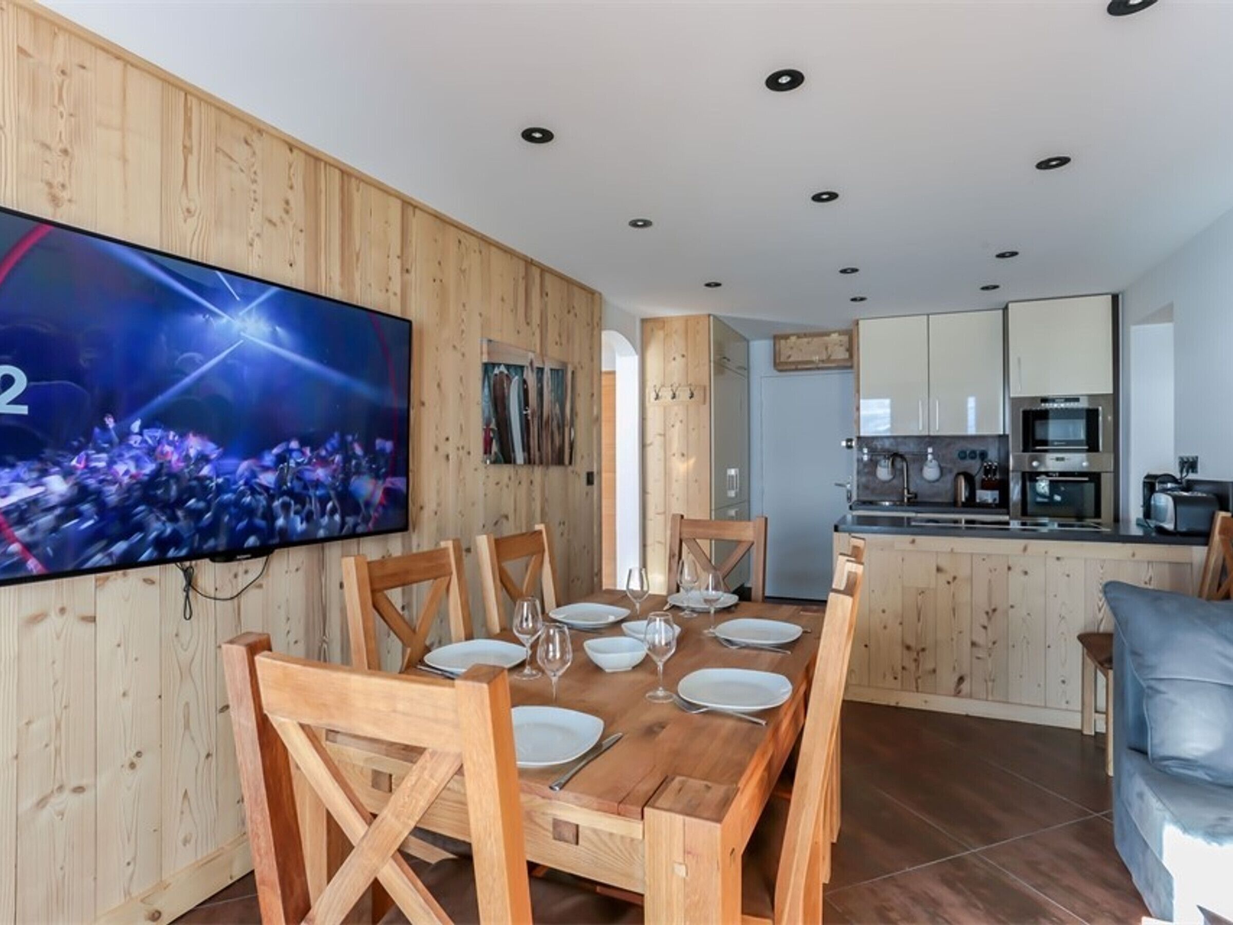 A dining area with a wooden table set for six, a TV mounted on the wall, and a kitchen in the background.