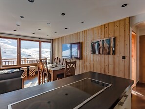 A modern dining area with a wooden wall, a large window offering a view of snow-covered mountains, and a flat-screen TV mounted on the wall. - Val Thorens: Stunning apt for 8, close to slopes and shops (Val Thorens)