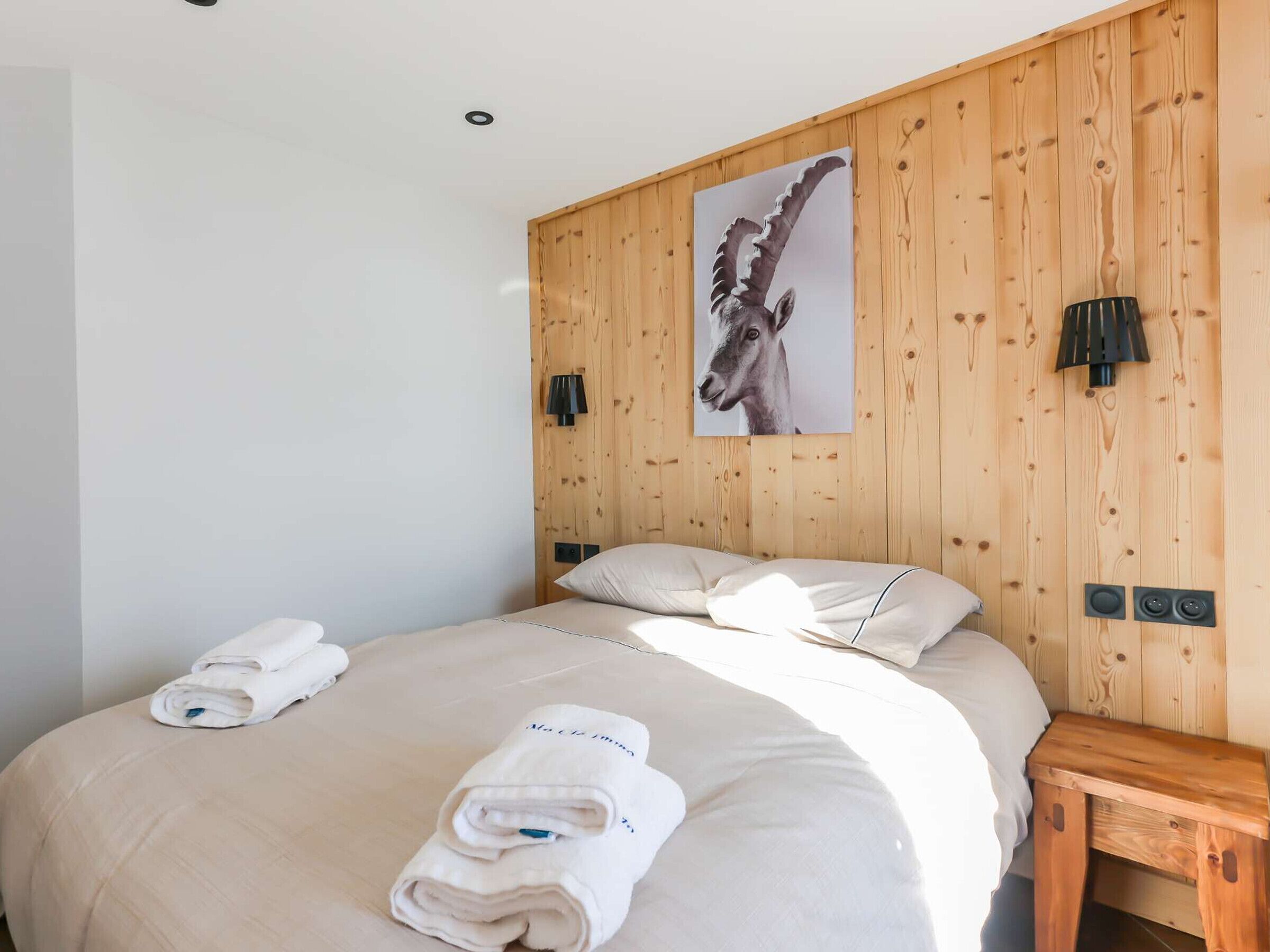 Een slaapkamer met een houten wand, een bed met witte lakens, een klein houten nachtkastje en een wandlamp.