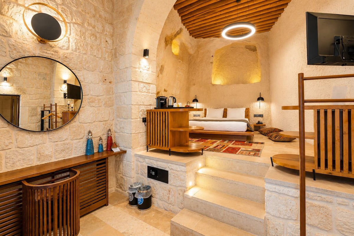 A stone-walled room with a bed, a wooden bench, a mirror, and a small table with decorative items.