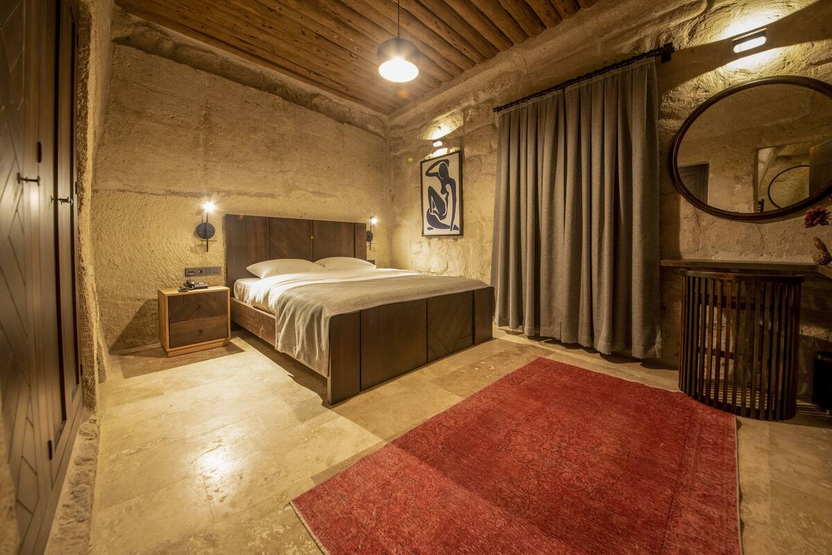 A bedroom with a bed, a nightstand, a red rug, a wooden ceiling, and a mirror.