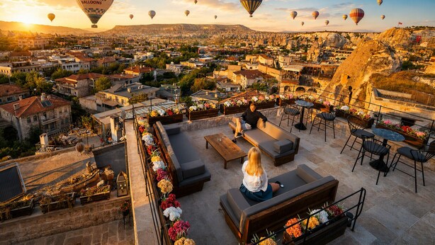 Una terraza en la azotea con asientos al aire libre, vistas a un paisaje urbano y globos aerostáticos en el cielo.