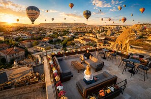 Una terraza en la azotea con asientos al aire libre, vistas a un paisaje urbano y globos aerostáticos en el cielo.