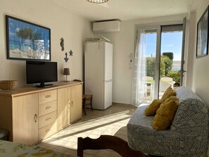 A room with a television on a wooden cabinet, a bed with patterned bedding, a small table with a lamp, and a view through a door to an outdoor area. - Charming 1-Bedroom Apartment with Private Terrace Near Thau Lagoon (Balaruc-les-Bains)