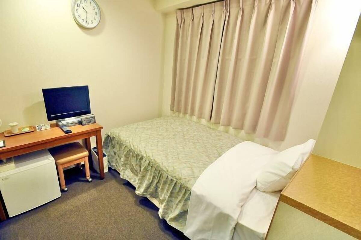 A small, neatly arranged bedroom with a single bed, a desk with a computer, a small refrigerator, and a clock on the wall.