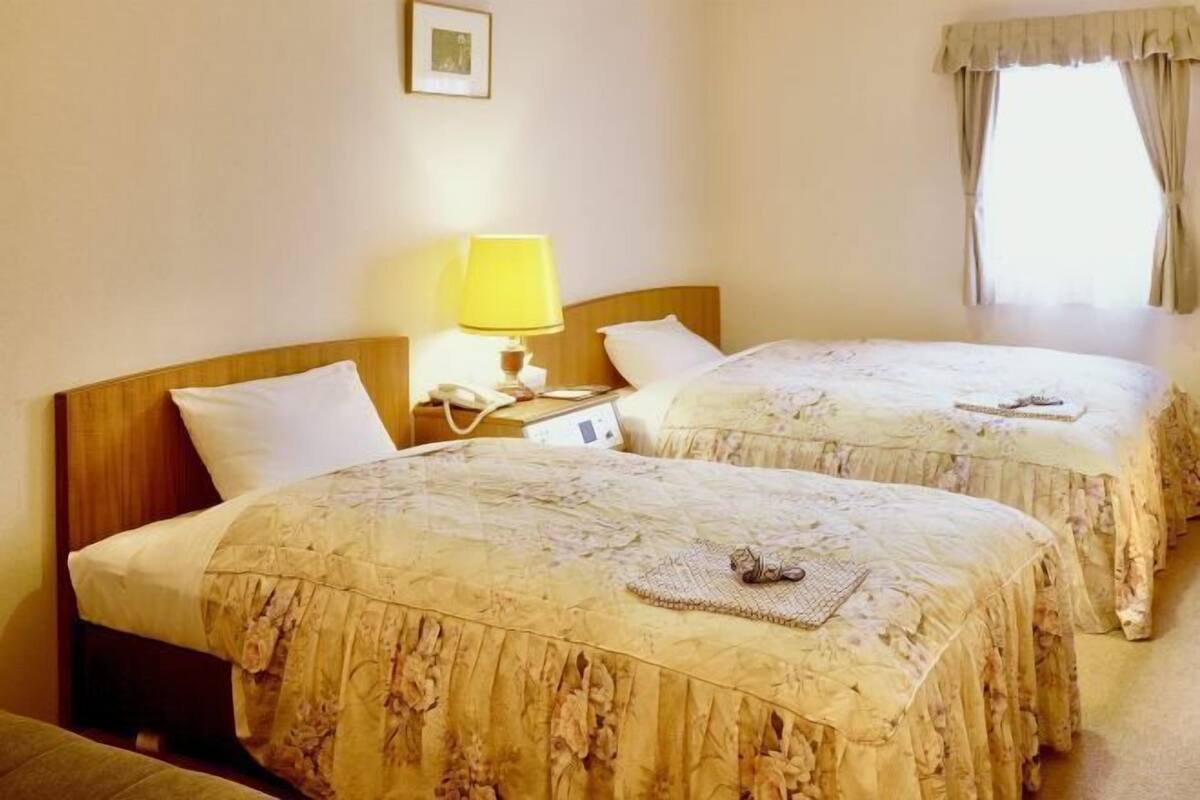 A hotel room with two beds, a bedside table with a lamp, a window with curtains, and a wall-mounted picture.