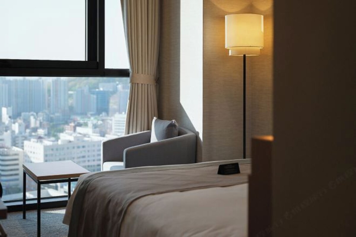 A hotel room with a bed, a chair, a small table, and a lamp. A cityscape is visible through the window.