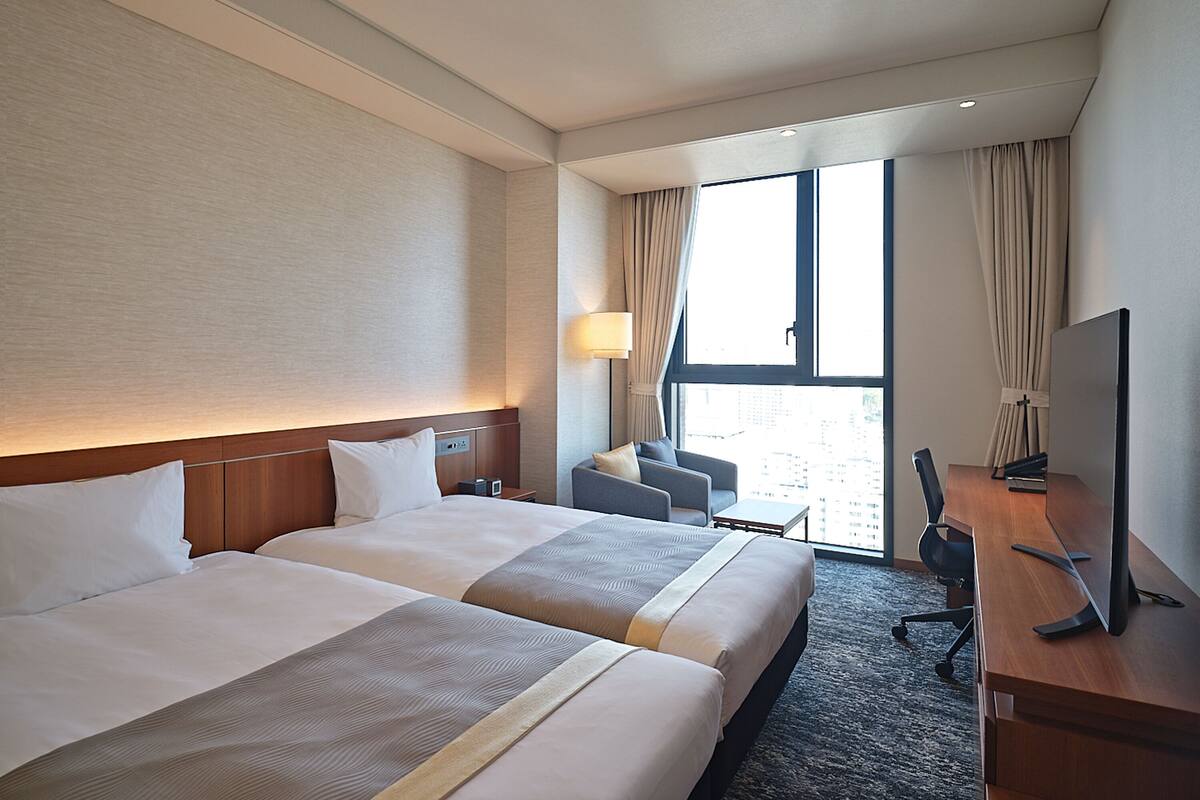 Une chambre d’hôtel avec deux lits, un bureau, une chaise, une télévision et une fenêtre avec des rideaux.