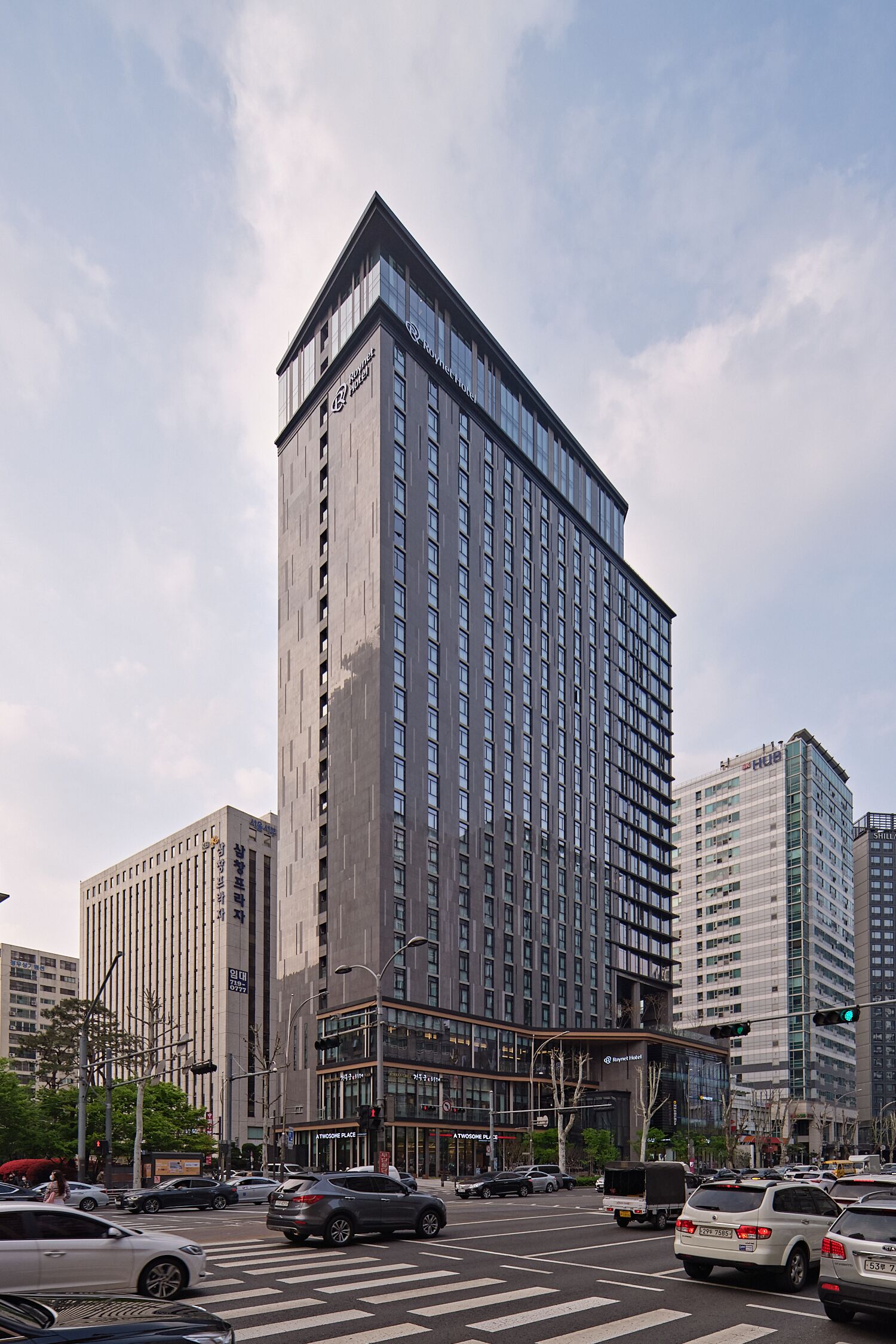 Un moderno edificio de gran altura con una fachada de vidrio destacada, probablemente un hotel o edificio de oficinas, ubicado en una concurrida intersección urbana con tráfico y farolas visibles.