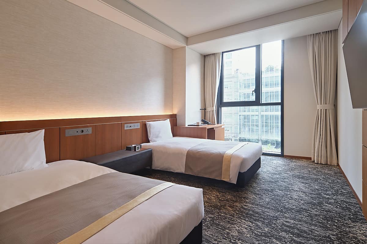 Une chambre d’hôtel avec deux lits, une grande fenêtre avec des rideaux, un téléviseur à écran plat et une tête de lit en bois.