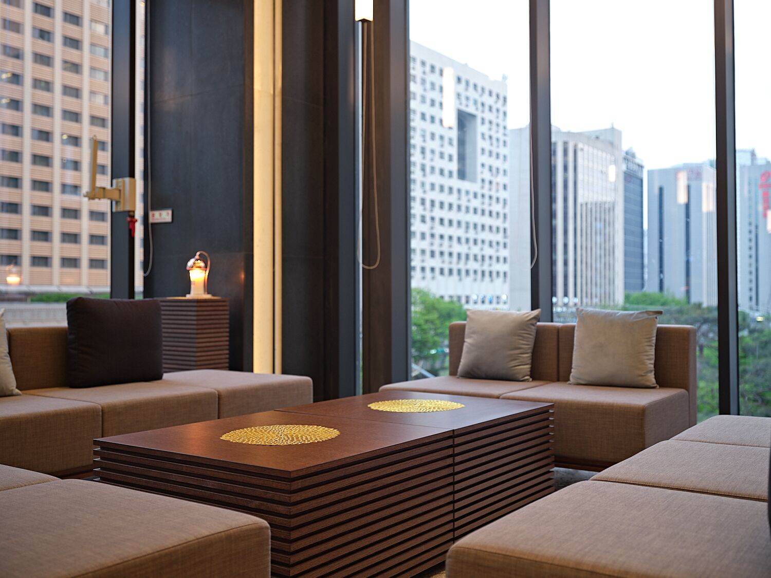 A modern interior with a sofa set, a wooden coffee table, and a view of tall buildings through large windows.