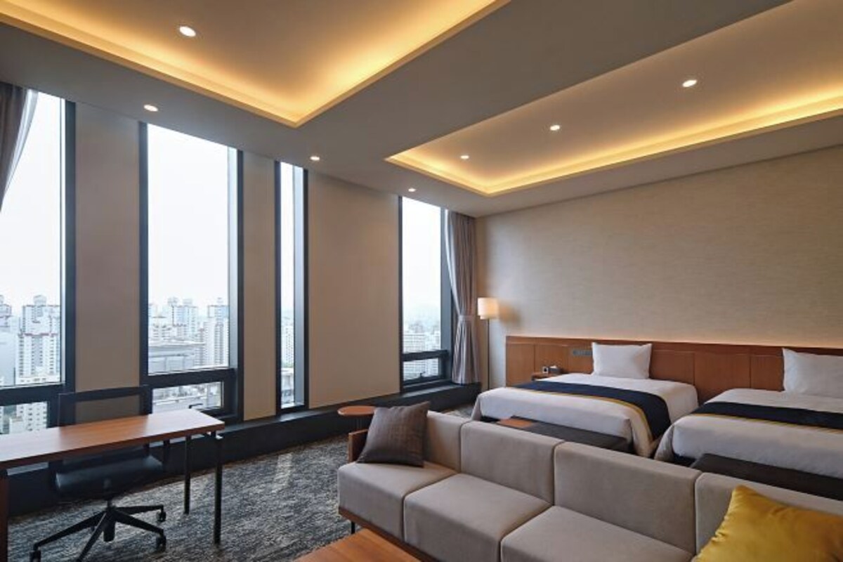 A hotel room with a sofa, two beds, a desk, and large windows offering a city view.