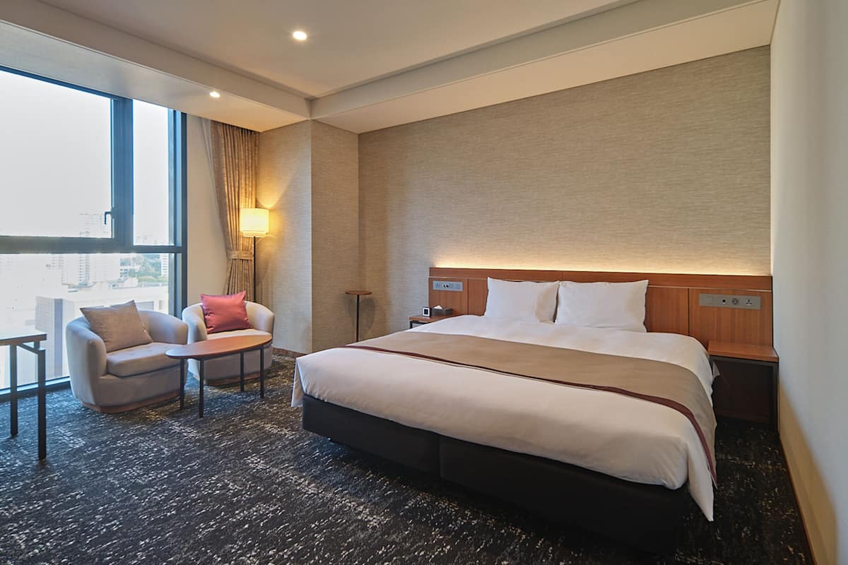Une chambre d’hôtel avec un grand lit, un bureau, une chaise et une fenêtre donnant sur des immeubles.